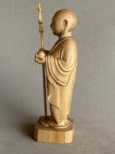 Light colored handcarved wood statue of Jizo as a man, holding a staff with 6 gold rings and in his other hand the Wish Fulfilling Gem, side view