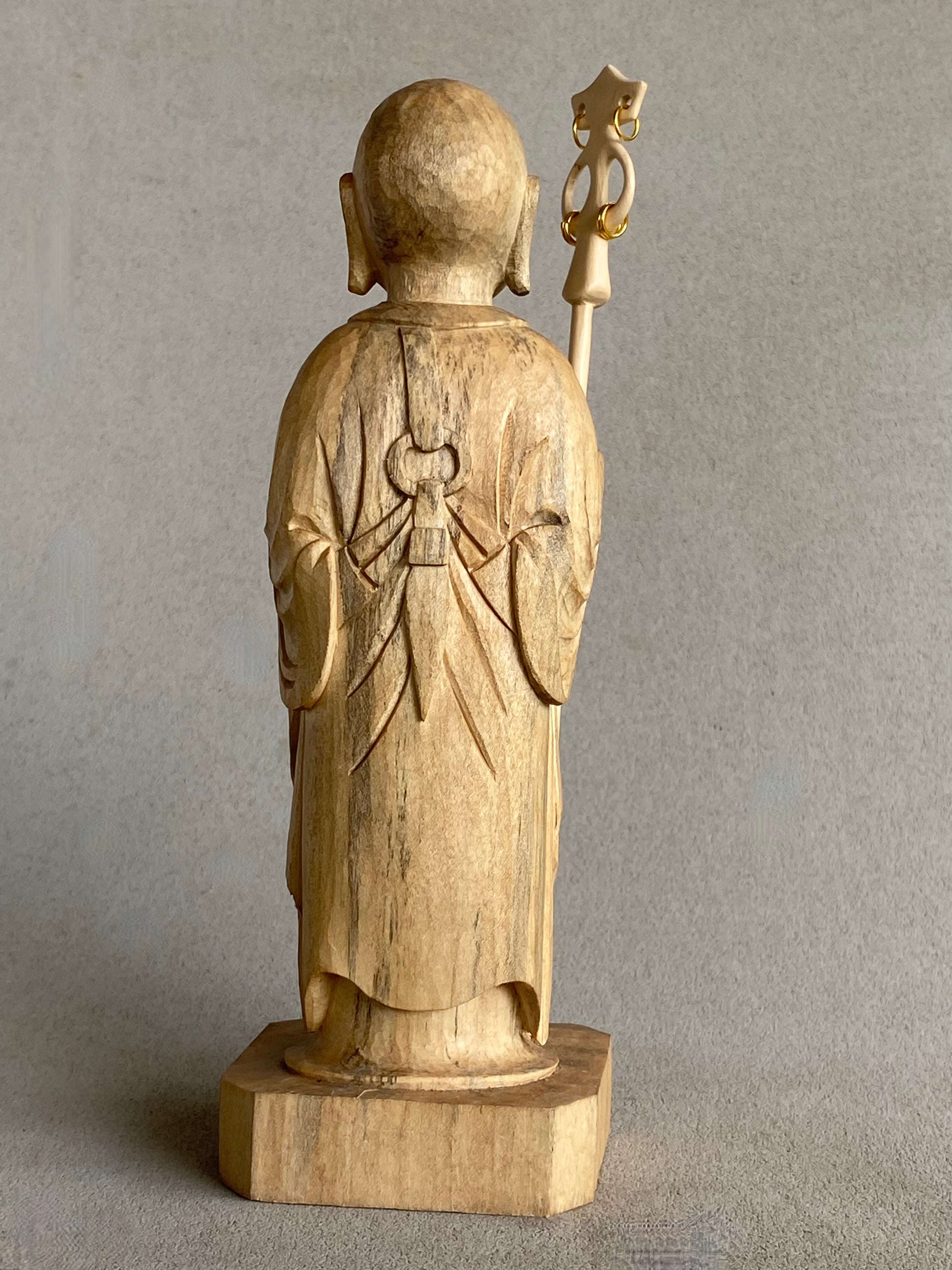 Light colored handcarved wood statue of Jizo as a man, holding a staff with 6 gold rings and in his other hand the Wish Fulfilling Gem, back view