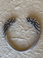 Load image into Gallery viewer, A silver-toned metal cuff bracelet with detailed dragon head with mane carvings on both ends, displayed on a textured cream hand made paper background.
