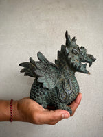 Load image into Gallery viewer, A hand holds a small dragon figurine with a textured, weathered green and bronze finish against a plain light gray background.

