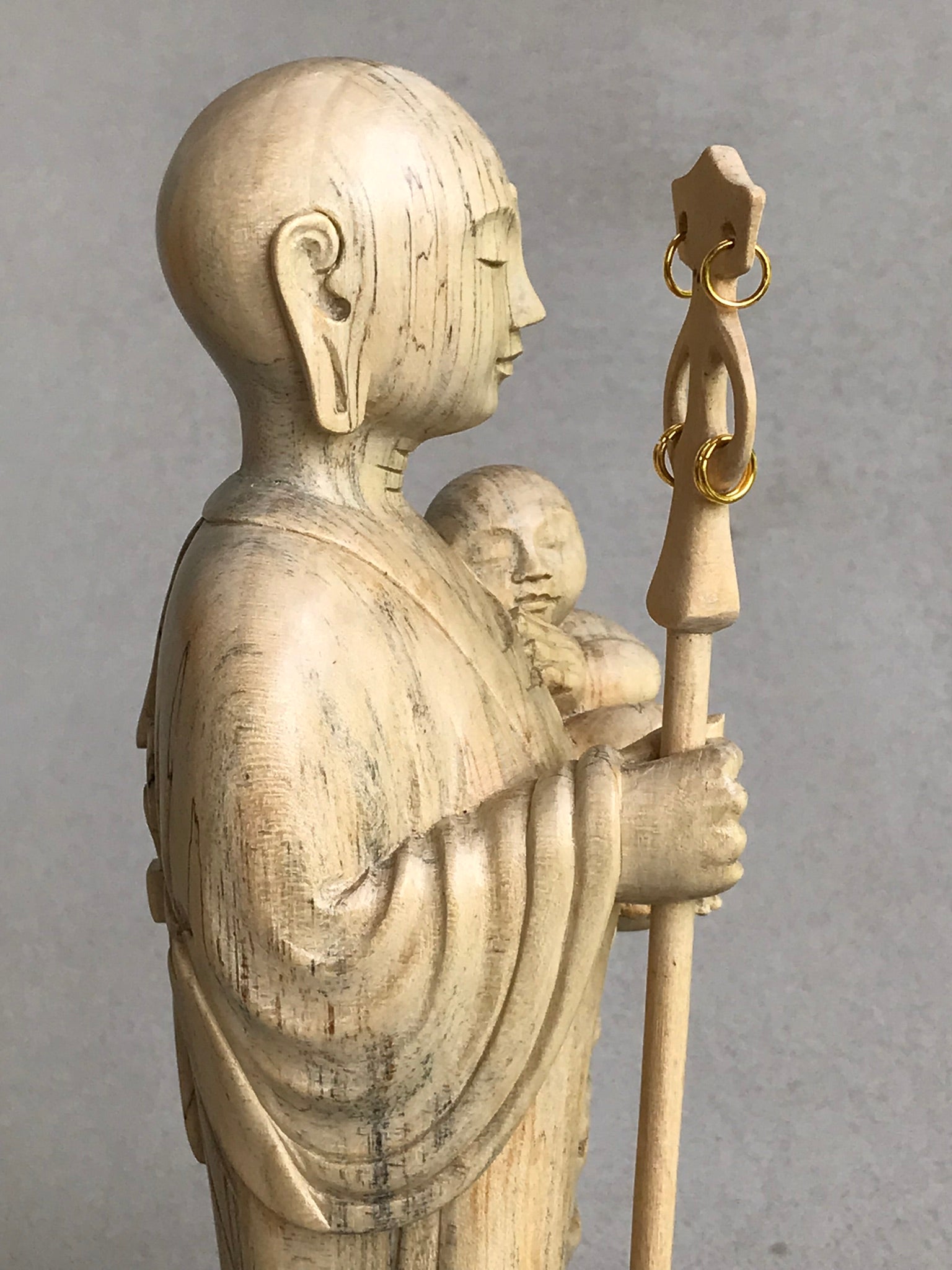 JIzo man standing, holding a staff and a baby, 25cm, 10 inches, color variation in wood from warm light brown to grey grain, close up view of right side.