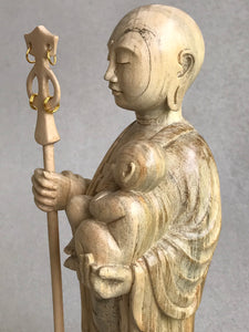 JIzo man standing, holding a staff and a baby, 25cm, 10 inches, color variation in wood from warm light brown to grey grain. close up view of left side